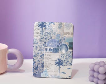 May include: A rectangular coaster with a blue and white coastal design featuring palm trees, seashells, and the text "Coastal Life" and "Mamm". The coaster is placed on a white surface with a pink mug and a purple bubble candle.