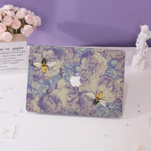 Könnte beinhalten: Eine Laptop-Hülle mit einem floralen Muster in Lila, Blau und Creme, verziert mit Bienen-Illustrationen. Die Hülle hat ein weißes Apple-Logo. Die Hülle ist ein schützendes Zubehör für einen Laptop.