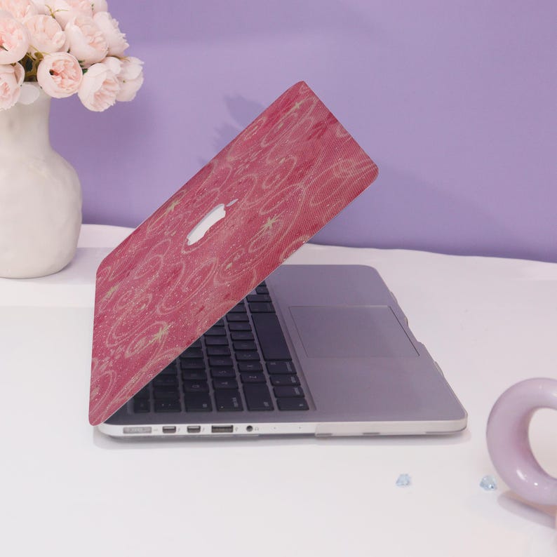 May include: A pink laptop case featuring a white Apple logo and a pattern of light yellow swirls and starbursts. The laptop is open, showing a black keyboard and a silver trackpad. The case is on a white surface.