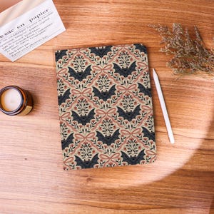 May include: A rectangular notebook featuring a repeating pattern of dark grey bats, pink bows, and small floral sprigs on a light beige background. The notebook is positioned next to a white stylus, a small candle in a glass jar, and a dried plant sprig.