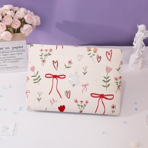May include: A white laptop with a floral and heart design. The design features red bows, pink flowers, and white hearts on a white background.