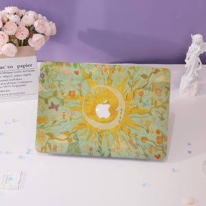 May include: A decorative laptop case featuring a sun and moon design with an Apple logo cutout. The case has a light green background with floral and butterfly accents. The sun and moon are yellow, and the overall aesthetic is whimsical.