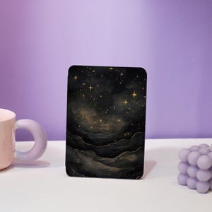 May include: A rectangular black and gold art print with a celestial design of stars and layered cloud-like shapes. The artwork has a watercolour effect and is displayed on a white surface. A pink mug and a lavender bubble candle are also in the image.