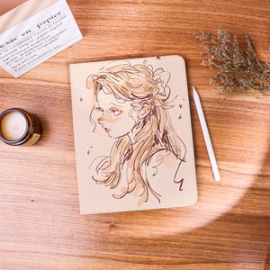 May include: A beige notebook with a sketch of a woman's face in brown ink. A white stylus rests beside the notebook on a wooden surface. A small candle in a brown jar and a paper bag are also visible.