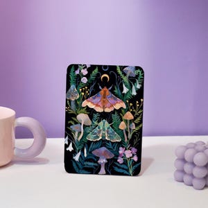 May include: A black background with a colourful illustration of moths and mushrooms. The moths are in the centre of the image and the mushrooms are around them. The illustration is surrounded by green leaves and flowers.