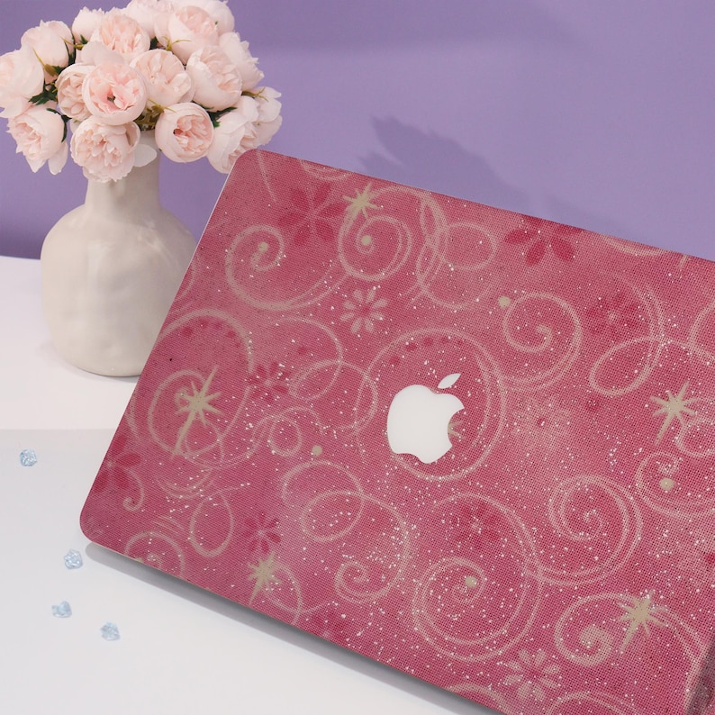 May include: Pink laptop case featuring a white Apple logo and a swirling floral design. The case has a glittery texture and a white floral pattern. A white vase with pink flowers is in the background.