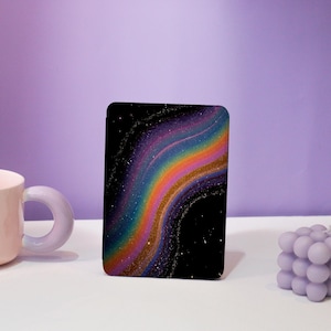 May include: A black rectangular case with a vibrant, swirling rainbow and star design. The design incorporates shades of purple, blue, green, yellow, orange, and gold. A light pink mug and a lavender cube candle are also present.