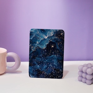Peut inclure: Un objet rectangulaire aux coins arrondis, orné d'un motif de galaxie bleu foncé et or. L'image présente un ciel étoilé avec des nuages tourbillonnants. L'objet est posé sur une surface blanche avec un fond violet.