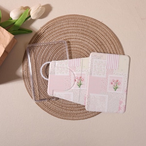 Może przedstawiać: Przezroczyste etui na telefon oraz różowo-białe etui na telefon z pasującą osłoną, z kwiatowym wzorem. Przedmioty ułożone są na tkanej macie, z bukietem białych tulipanów w tle.