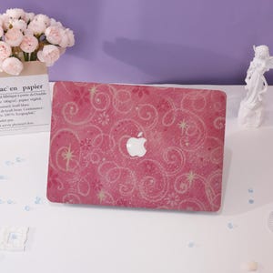 Può includere: Custodia rosa per laptop con logo Apple bianco. La custodia presenta un design floreale a spirale con accenti glitterati. La custodia è su una superficie bianca con un bouquet di fiori e una piccola statua sullo sfondo.