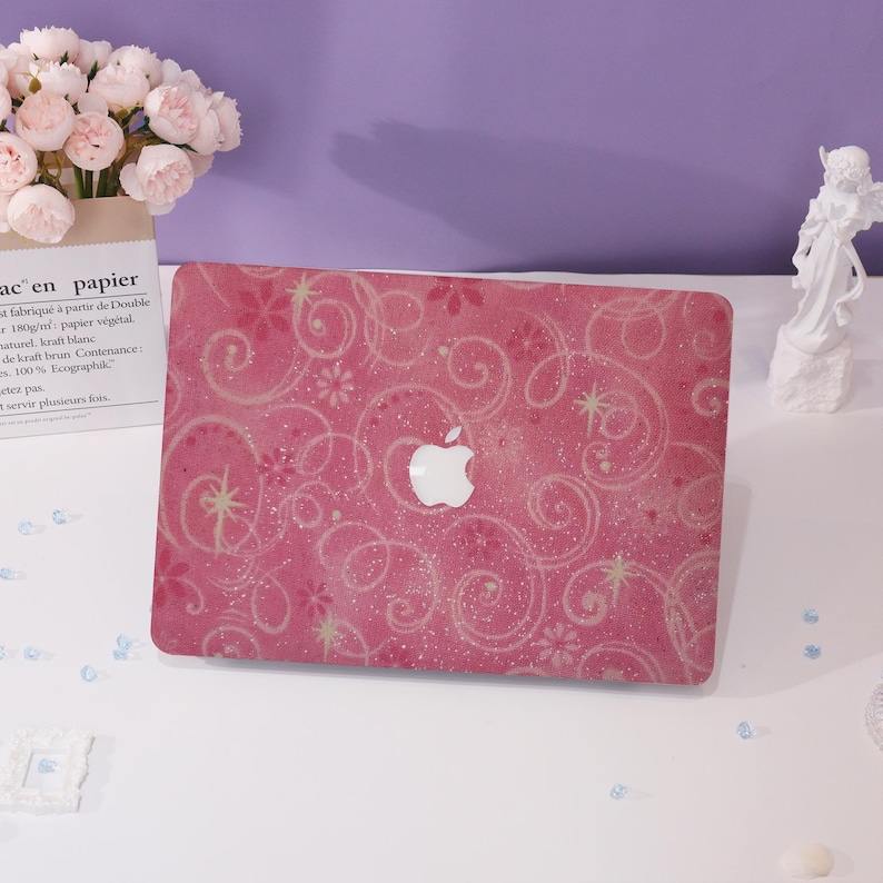 May include: Pink laptop case with a white Apple logo. The case has a swirling floral design with glitter accents. The case is on a white surface with a bouquet of flowers and a small statue in the background.