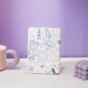 Puede incluir: Una funda de teléfono blanca con un patrón de garabatos azul y blanco. El patrón incluye botas de vaquero, estrellas, corazones, conchas marinas y las palabras "Howdy", "Yee Haw" y "Lucky".