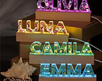 May include: Illuminated name signs with floral designs. The names "OLIVIA", "LUNA", "CAMILA", and "EMMA" are displayed in colorful, glowing letters. Each name is set on a wooden base, creating a decorative and personalized lighting effect.