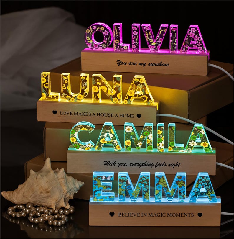 May include: Illuminated name signs with floral designs. The names Olivia, Luna, Camila, and Emma are displayed in colorful, glowing letters. Each name sits on a wooden base with inspirational quotes. A seashell and pearls are in the foreground.