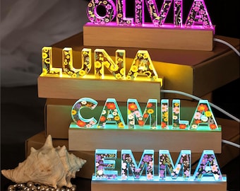 May include: Four custom name signs featuring the names OLIVIA, LUNA, CAMILA, and EMMA. Each sign consists of clear resin letters embedded with colorful dried flowers, mounted on a rectangular wooden base that illuminates the letters from below.