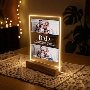 May include: A rectangular LED photo frame with a wooden base. The frame displays two family photos and the word "DAD" with the text "TO THE WORLD YOU ARE A DAD BUT TO OUR FAMILY YOU ARE THE WORLD". The frame is illuminated with a warm, yellow light.
