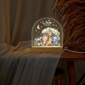 May include: A wooden night light with a clear acrylic panel featuring a whimsical scene of a zebra, lion, giraffe, and elephant. The panel is illuminated with a warm white light. The night light is personalized with the name "Liam" and the date "15.06.2021".
