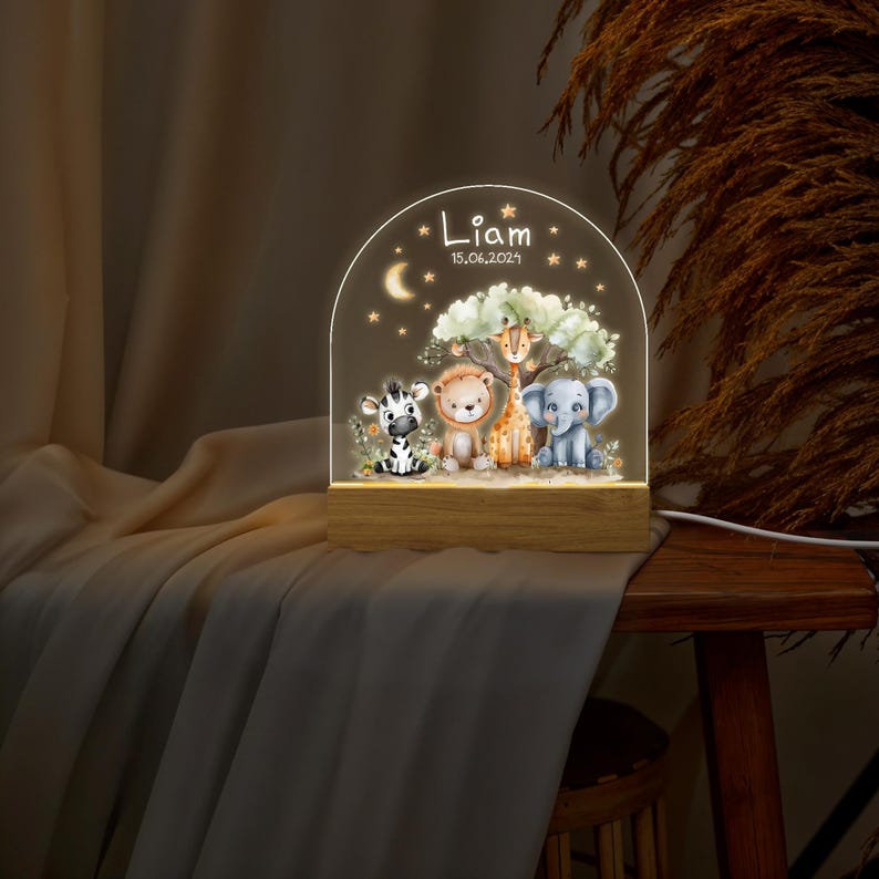May include: A wooden night light with a clear acrylic panel featuring a whimsical scene of a zebra, lion, giraffe, and elephant. The panel is illuminated with a warm white light. The night light is personalized with the name "Liam" and the date "15.06.2021".