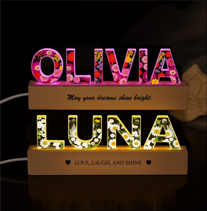 May include: Illuminated floral name signs, featuring the names "OLIVIA" and "LUNA" in bright, colorful letters. The signs are set on wooden bases, with the text "May your dreams shine bright" and "LOVE, LAUGH, AND SHINE".