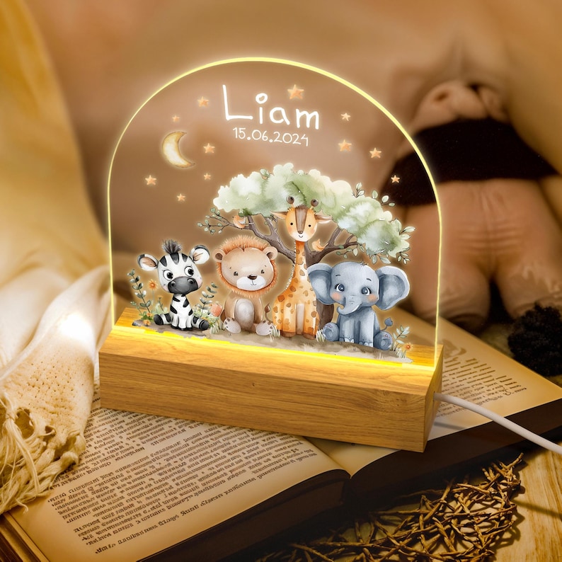 May include: A wooden night light with a clear acrylic dome featuring a whimsical jungle scene with a zebra, lion, giraffe, and elephant. The dome is illuminated with a warm white light and the base is engraved with the name "Liam" and the date "15.06.2024".
