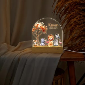 May include: A personalised night light with a wooden base and a clear acrylic dome. The dome features a colourful illustration of a jungle scene with a lion, zebra, hippopotamus, tiger, and flamingo. The text "Kevin 10.06.2024" is printed on the dome.