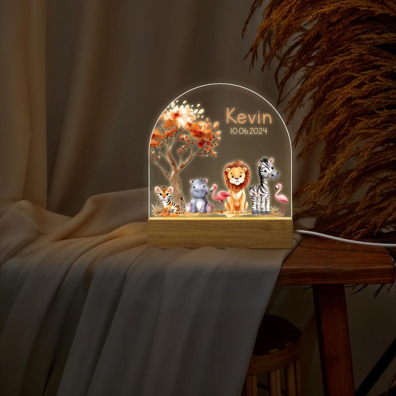 May include: A personalised night light with a wooden base and a clear acrylic dome. The dome features a colourful illustration of a jungle scene with a lion, zebra, hippopotamus, tiger, and flamingo. The text "Kevin 10.06.2024" is printed on the dome.