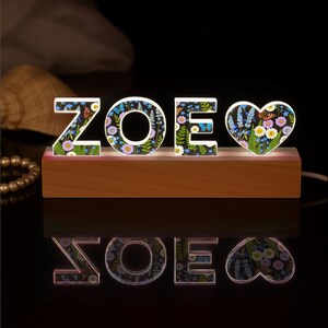 May include: A wooden base supports the illuminated letters "ZOE" and a heart-shaped design. The letters and heart are decorated with a floral pattern featuring butterflies and various flowers. The light shines upwards, creating a reflection.