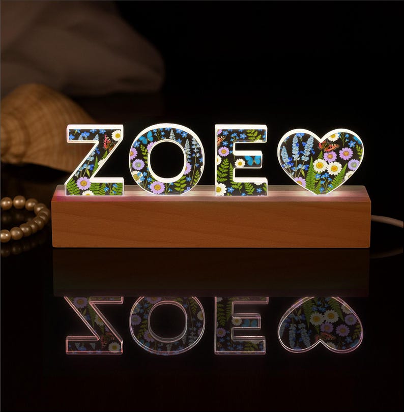 May include: A wooden base supports the illuminated letters "ZOE" and a heart-shaped design. The letters and heart are decorated with a floral pattern featuring butterflies and various flowers. The light shines upwards, creating a reflection.