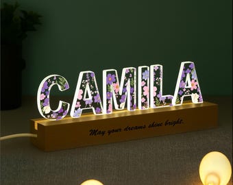 May include: A lighted sign spelling "CAMILA" with floral designs inside the letters. The sign sits on a wooden base with the phrase "May your dreams shine bright." The letters are illuminated with a soft glow.