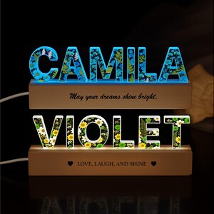 May include: Two illuminated wooden name signs. The top sign spells "CAMILA" in blue-lit letters filled with floral designs. The bottom sign spells "VIOLET" in white and yellow floral letters. Text on the signs reads "May your dreams shine bright" and "LOVE, LAUGH, AND SHINE."