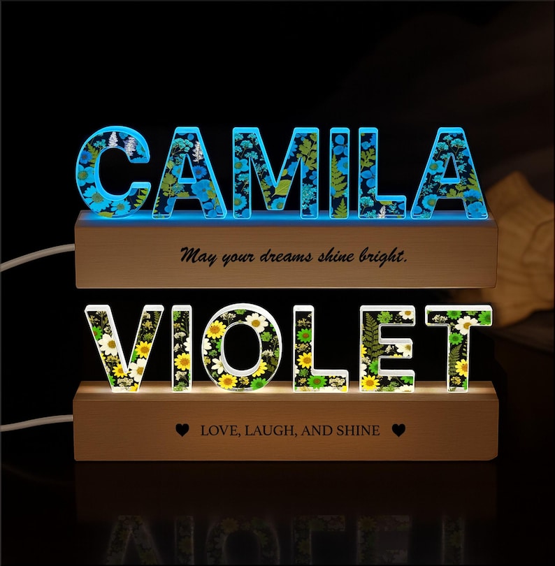 May include: Two illuminated wooden name signs. The top sign spells "CAMILA" in blue-lit letters filled with floral designs. The bottom sign spells "VIOLET" in white and yellow floral letters. Text on the signs reads "May your dreams shine bright" and "LOVE, LAUGH, AND SHINE."