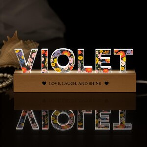 May include: A decorative illuminated sign spelling "VIOLET" with colorful floral patterns. The letters are set on a wooden base with the text "Love, Laugh, and Shine". The sign is lit from within, creating a warm glow.