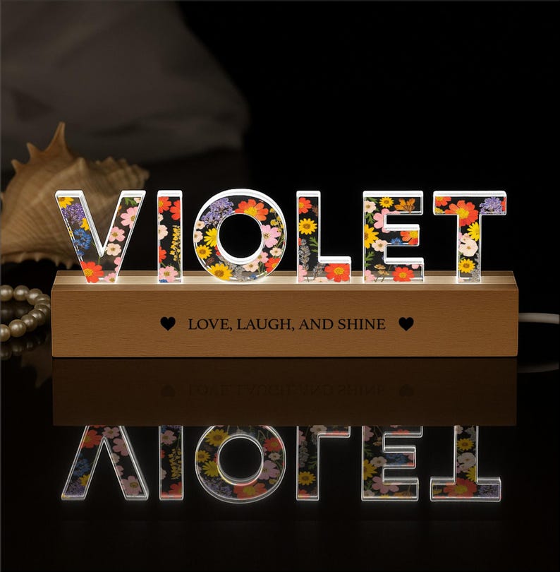 May include: A decorative illuminated sign spelling "VIOLET" with colorful floral patterns. The letters are set on a wooden base with the text "Love, Laugh, and Shine". The sign is lit from within, creating a warm glow.