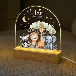 Può includere: Una luce notturna in legno con un pannello in acrilico trasparente che presenta un'illustrazione fantasiosa di una zebra, un leone, una giraffa e un elefante in un ambiente di giungla. Il pannello è illuminato da luci LED bianco caldo. Il pannello in acrilico include anche il nome "Liam" e la data "15.06.2024".