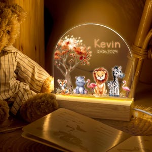 May include: A personalised night light with a wooden base and a clear acrylic panel featuring a jungle scene with a tiger, hippopotamus, lion, zebra, and flamingos. The panel is illuminated with warm white LED lights. The text "Kevin 10.06.2024" is printed on the panel.