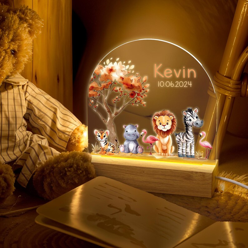May include: A personalised night light with a wooden base and a clear acrylic panel featuring a jungle scene with a tiger, hippopotamus, lion, zebra, and flamingos. The panel is illuminated with warm white LED lights. The text "Kevin 10.06.2024" is printed on the panel.