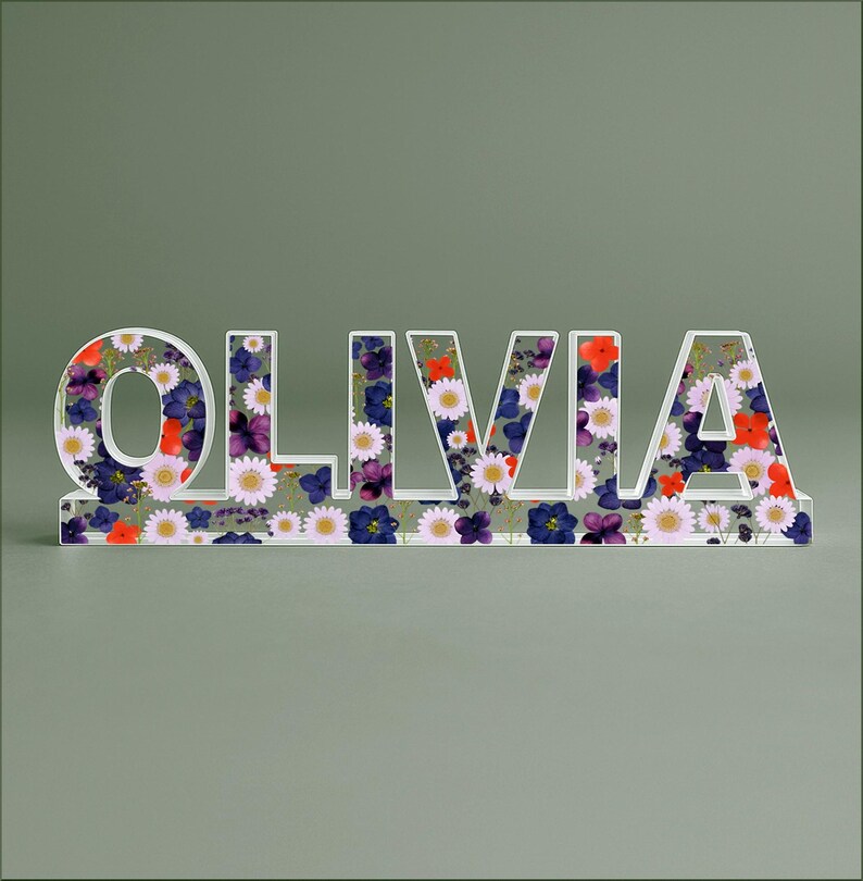 May include: A decorative sign spelling "OLIVIA" in white, with a floral pattern of red, purple, and white flowers. The sign is made of a clear material and is designed to stand upright.