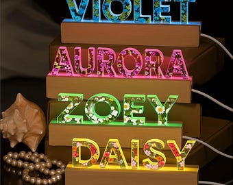 May include: Illuminated name signs, each featuring a different name: Violet, Aurora, Zoey, and Daisy. The letters are filled with floral patterns and sit atop wooden bases. Each name glows with a different color: blue, pink, green, and yellow.