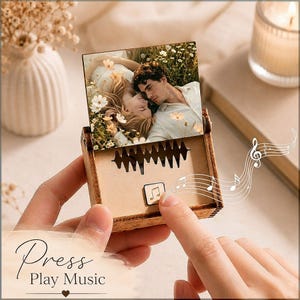 May include: A square wooden music box featuring a colour photograph of a couple lying in a field of flowers. The box has a cutout sound wave design on the front and a small square button with a musical note icon. The text Press Play Music is visible.