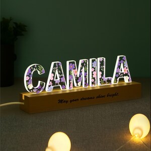 May include: A lighted sign spelling "CAMILA" with floral designs inside the letters. The sign sits on a wooden base with the phrase "May your dreams shine bright." The letters are illuminated with a soft glow.