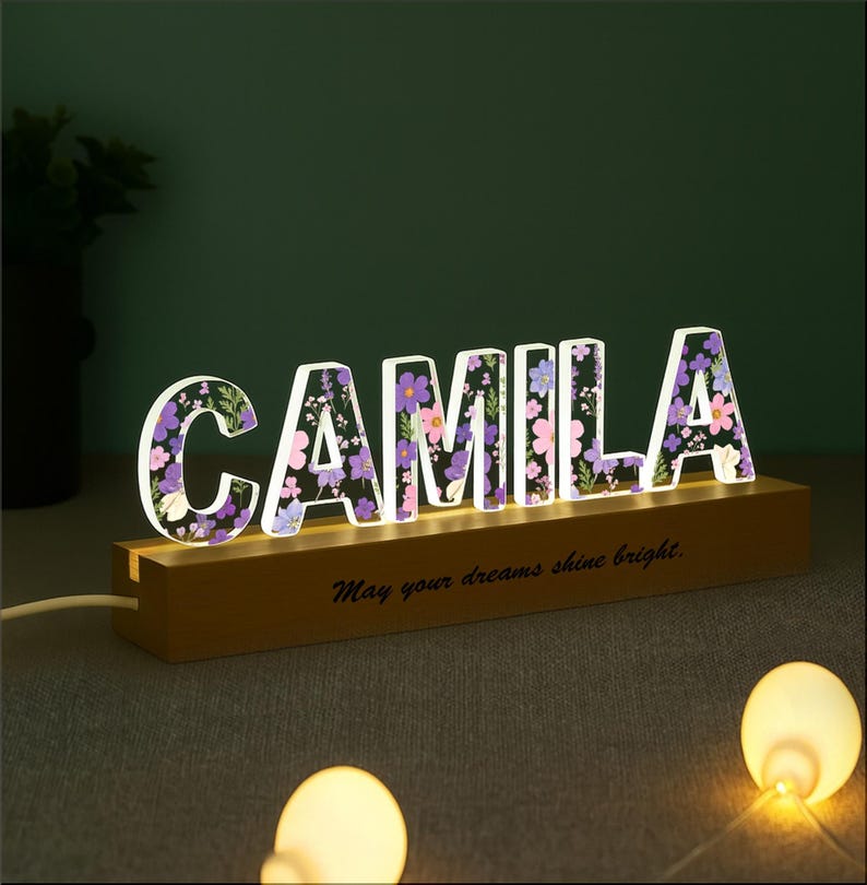 May include: A lighted sign spelling "CAMILA" with floral designs inside the letters. The sign sits on a wooden base with the phrase "May your dreams shine bright." The letters are illuminated with a soft glow.