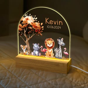 May include: A wooden night light with a clear acrylic insert featuring a jungle scene with a lion, zebra, hippopotamus, tiger, and flamingos. The acrylic insert is illuminated with a warm white light. The night light is personalised with the name "Kevin" and the date "10.06.2024".
