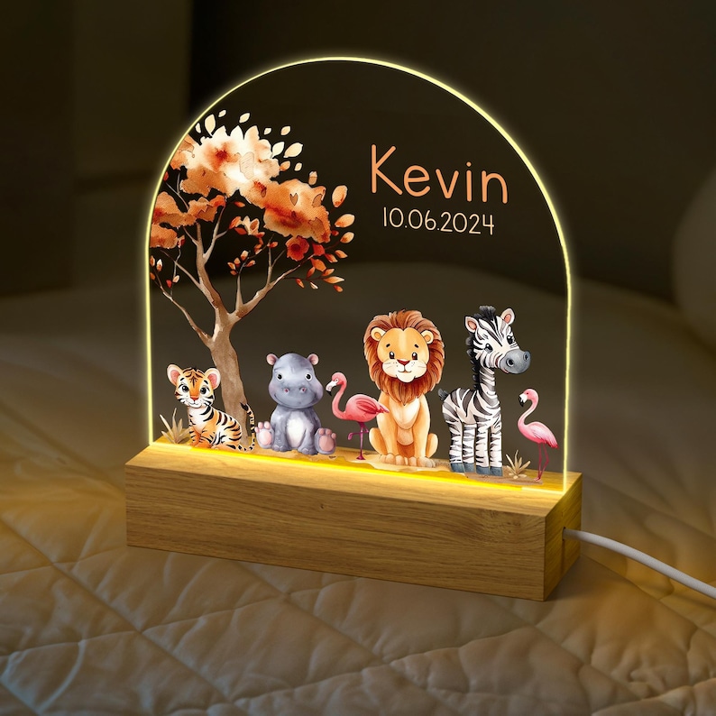 May include: A wooden night light with a clear acrylic insert featuring a jungle scene with a lion, zebra, hippopotamus, tiger, and flamingos. The acrylic insert is illuminated with a warm white light. The night light is personalised with the name "Kevin" and the date "10.06.2024".