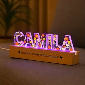 May include: A lighted sign spelling "CAMILA" with floral designs inside the letters. The letters are lit with a purple glow and sit on a wooden base. The base has the text "LOVE YOU TO THE MOON AND BACK".