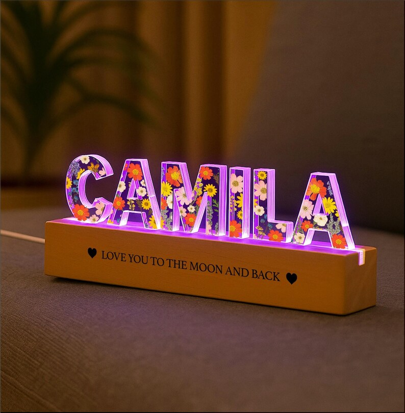 May include: A lighted sign spelling "CAMILA" with floral designs inside the letters. The letters are lit with a purple glow and sit on a wooden base. The base has the text "LOVE YOU TO THE MOON AND BACK".