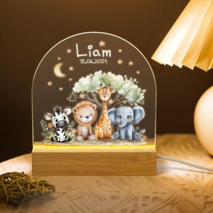 May include: A wooden night light with a clear acrylic panel featuring a whimsical jungle scene with a zebra, lion, giraffe, and elephant. The panel is illuminated with a warm white light. The base of the night light is engraved with the name "Liam" and the date "15.06.2024".