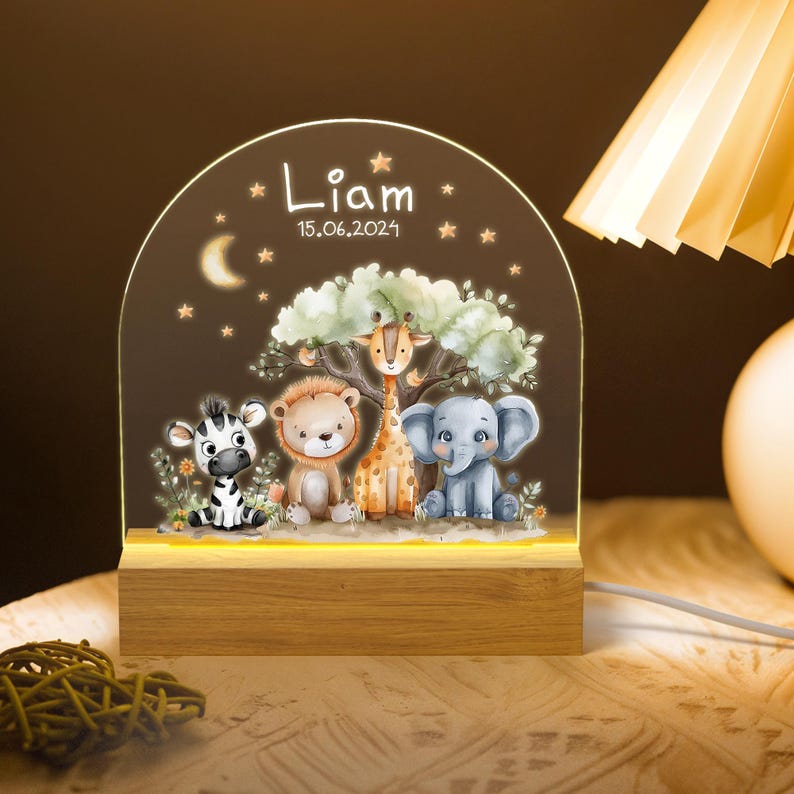 May include: A wooden night light with a clear acrylic panel featuring a whimsical jungle scene with a zebra, lion, giraffe, and elephant. The panel is illuminated with a warm white light. The base of the night light is engraved with the name "Liam" and the date "15.06.2024".