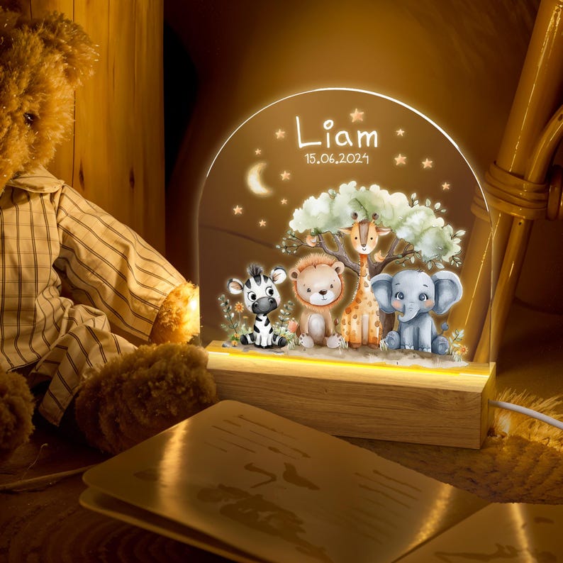 May include: A personalized night light with a wooden base and a clear acrylic panel featuring a whimsical jungle scene with a zebra, lion, giraffe, and elephant. The panel is illuminated with warm white LED lights. The name "Liam" and the date "15.06.2024" are printed on the panel.