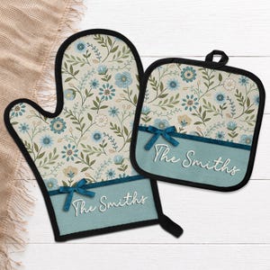 May include: Oven mitt and pot holder set with a floral pattern in blue, green, and beige. The set features a teal band with the text "The Smiths" and a teal bow. The edges are trimmed in black.