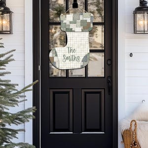 Puede incluir: Adorno de puerta de calcetín navideño de madera con diseño de patchwork en tonos verdes, blancos y beige. Personalizado con el nombre "The Smiths". Cuelga de una puerta negra.